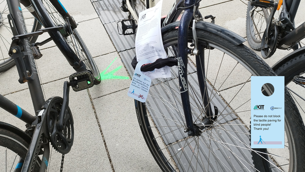 Vor einem Gebäude stehen Fahrräder, wob ein Rad steht auf der Leitlinie abgestellt ist. Ein Symbol  auf der Erde (Person mit Blindenstock) weist auf die Leitlinie hin. Der Sensibilisierungsanhänger "bitte Leitlinie freihalten"  ist abgebildet.